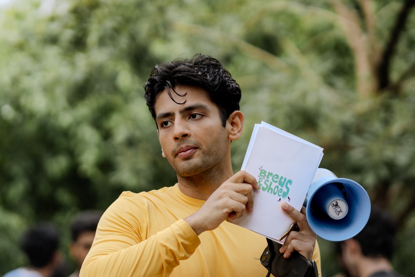 An organiser holding a hand-lettered 'Brews & Shoes' card and a megaphone, briefing the group before the run begins