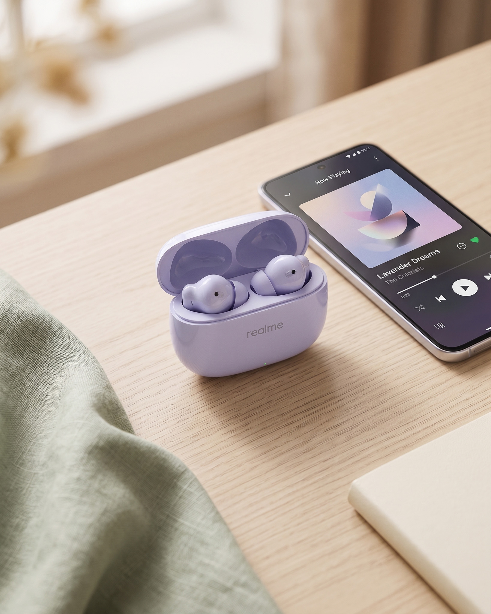 realme Buds T310 charging case open on a wooden desk, earbuds visible, next to a smartphone playing music