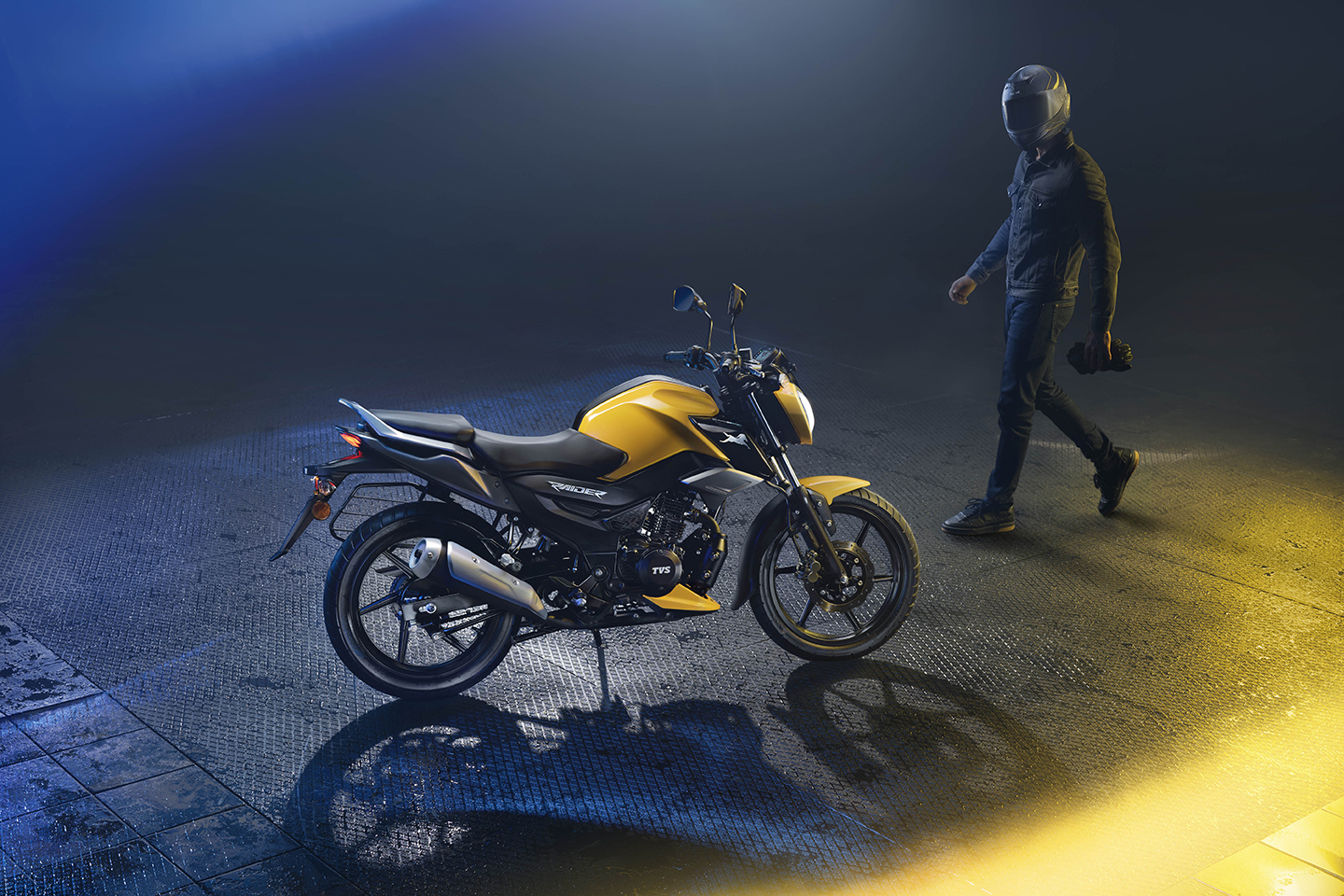 A rider walking back toward a parked yellow TVS Raider, helmet in hand, under dramatic studio lighting